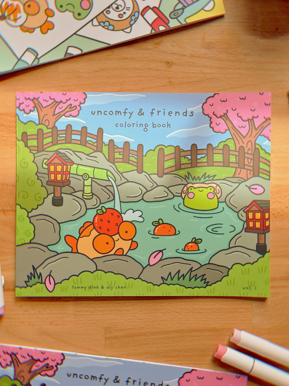 A coloring book titled 'Uncomfy & Friends' with illustrations of colorful characters and everyday scenes, placed on a wooden surface with a pen beside it.