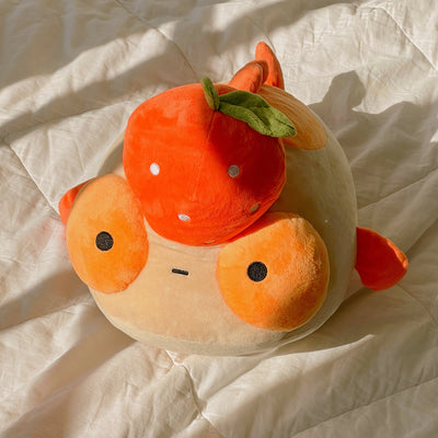 A plush orange goldfish with a tangerine on it's head, sitting in a sunbeam on a white quilt. 