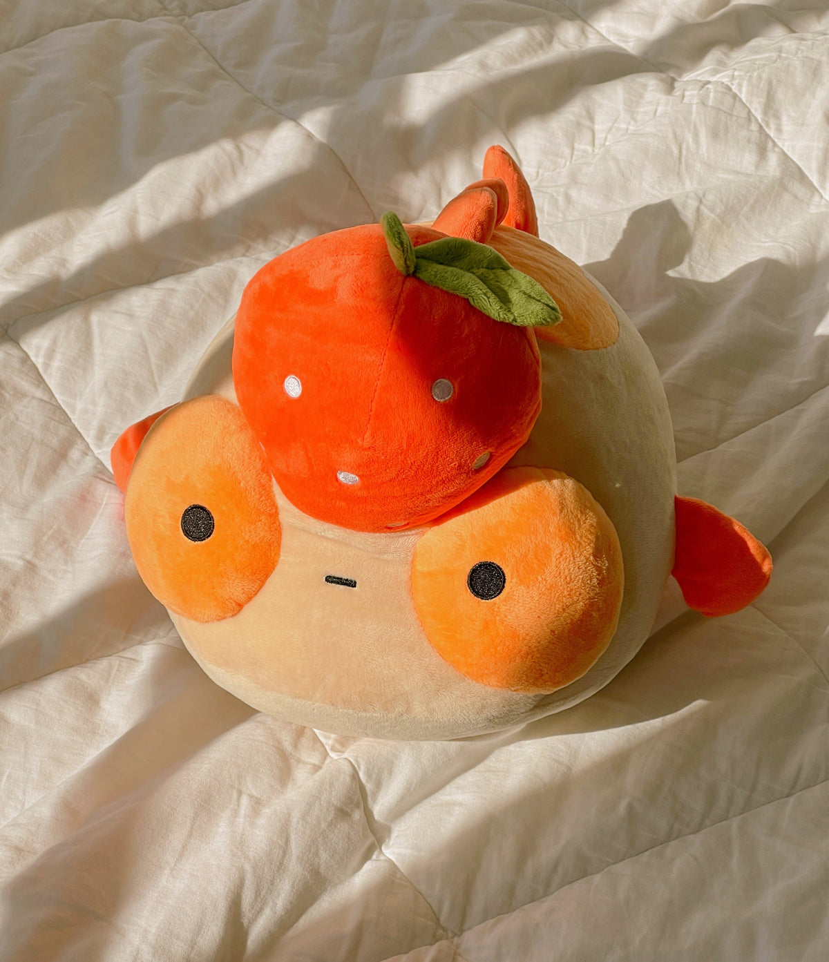 A plush orange goldfish with a tangerine on it's head, sitting in a sunbeam on a white quilt. 