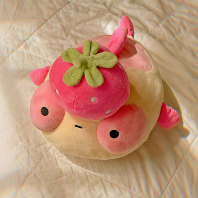 A plush pink goldfish with a strawberry on it's head, sitting on a white quilt. 