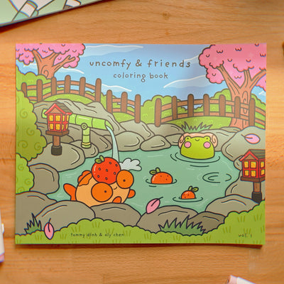 A coloring book titled 'Uncomfy & Friends' with illustrations of colorful characters and everyday scenes, placed on a wooden surface with a pen beside it.